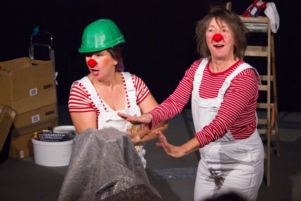 Zwei Clownetten bei einer Aufführung. Die Frau auf der linken Seite trägt einen grünen Bauarbeiterhelm. Beide Frauen tragen rote Shirts, darüber weiße Latzhosen und rote Nasen.