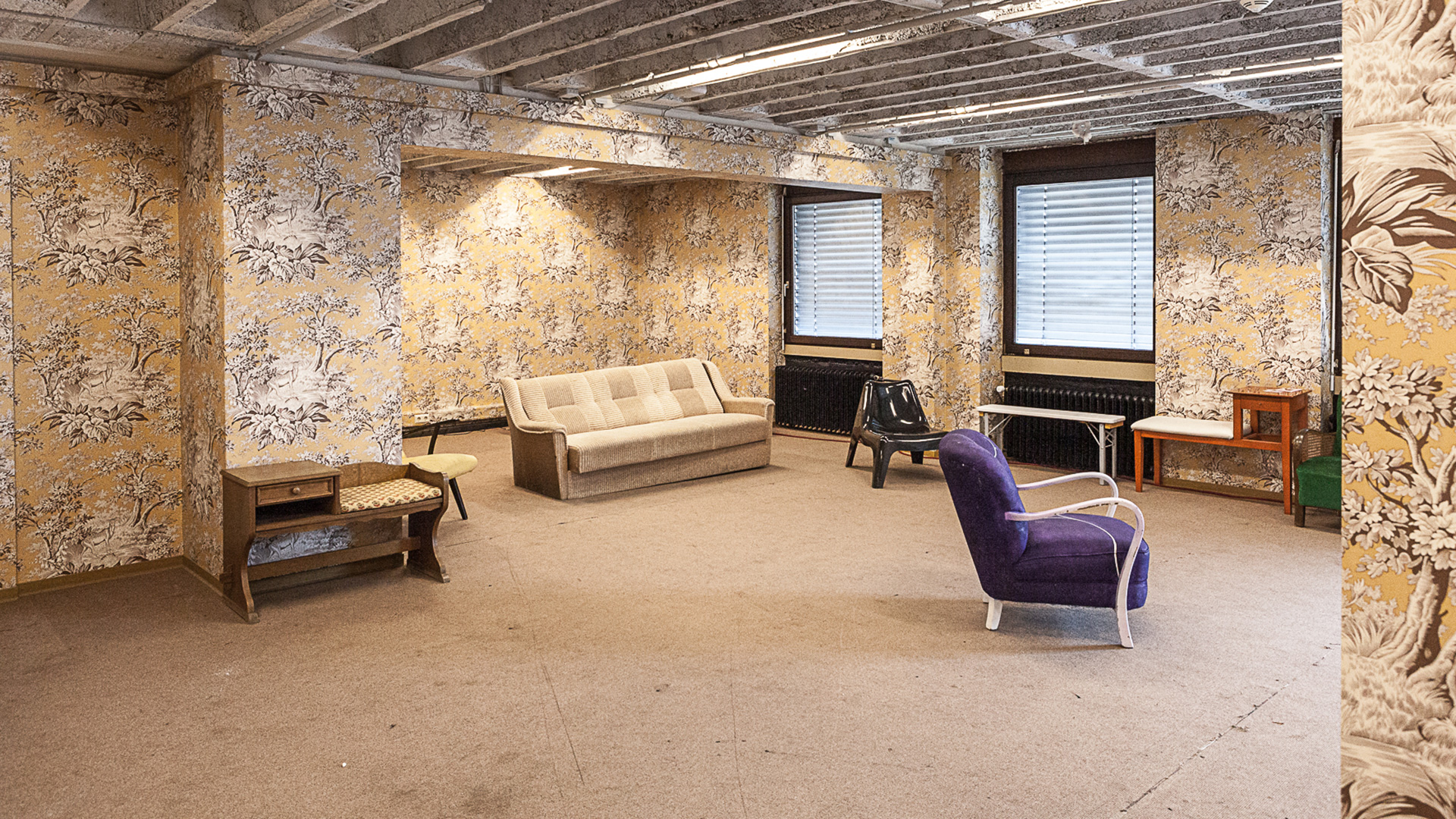 Das Bild zeigt einen großen Raum mit einer auffällig gemusterten Blumentapete und Möbel wie z.B. einem Sessel und einem Sofa.